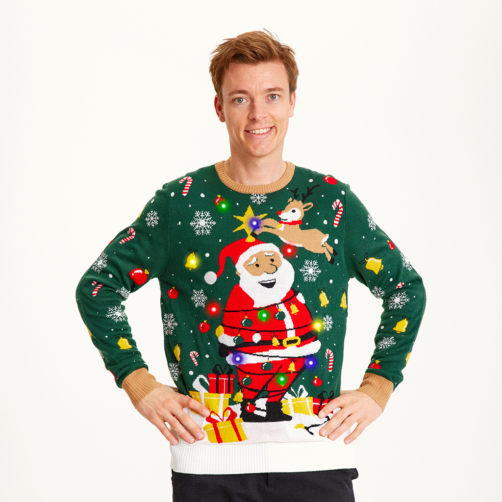 Un homme souriant, les mains le long du corps, portant un pull de Noël vert et blanc avec le Père Noël et des lumières.